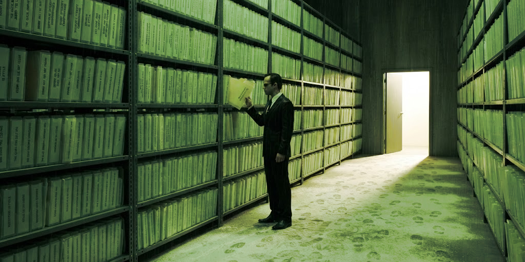 A package-repository stockroom in the green of a rain-lit night: steel shelving receding into the dark, every shelf holding translucent folders standing on edge like books, all of them empty, each labelled not with a name but with a sentence. A dusting of white flour on the concrete floor holds hundreds of identical shoe prints, all one size, walking every aisle. Halfway down the nearest aisle a man in a dark suit, sunglasses and an earpiece has stopped with his head turned, reading a label. At the far end a door stands open onto the outside and throws a slab of ordinary daylight into the green.